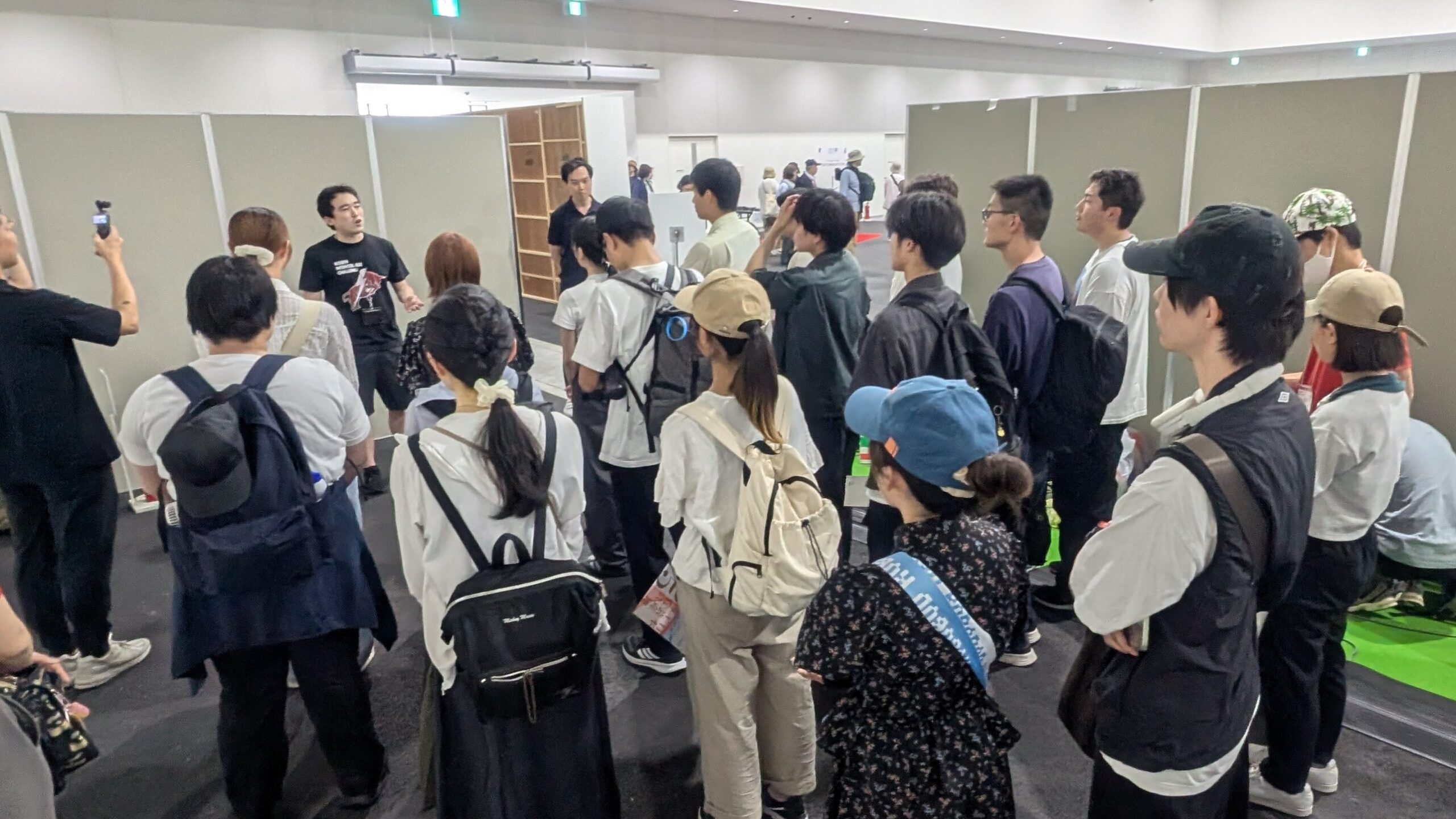 大阪関西万博で実施した「高専生の万博探求フィールドワーク」の一コマ。フィールドワークに出発する前の25名の高専生に対して、主催者であるSMBC日興証券の磯野さんと協賛企業である旭化成の橋詰さんが、激励の言葉をかけている写真。その場にいる高専生25名は二人の話を真面目に話を聞いている様子が伺える。6月中旬というのに30度を超える気温のため、日よけ対策の帽子をかぶっている学生が目立つ。